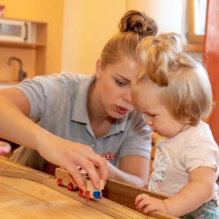 Betreuerin im Babymio spielt mit Kleinkind und Holzlokomotive