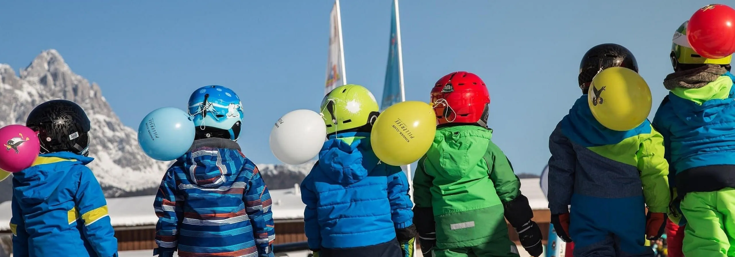 Kinder im Skianzug mit Helmen und Luftballons vor verschneiten Kitzbüheler